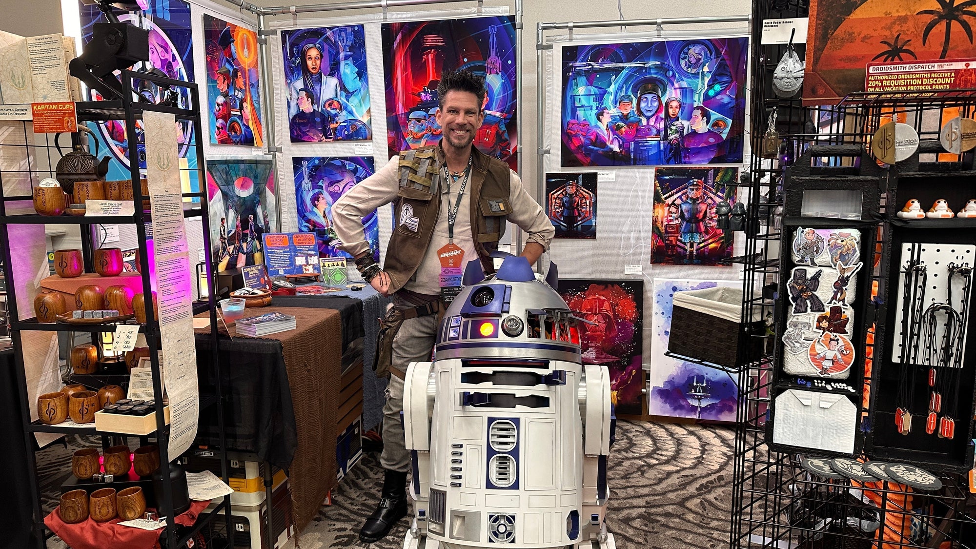 Nelson Ruger of 5th & Rugged at the Halcy-Con 2025 booth, surrounded by vivid Star Wars–inspired artwork and standing beside an R2-D2 droid.