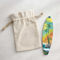 Hawaiian Holiday surfboard ornament displayed beside a natural canvas gift bag, hand-painted with island scenery