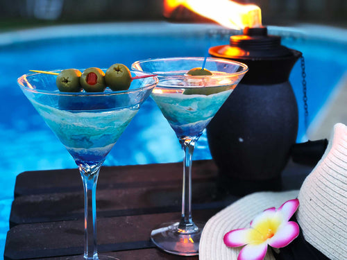 Pair of ocean wave martini glasses with cocktails and olive garnishes. Sitting beside a pool with a tropical flower hat and tiki fire bowl.
