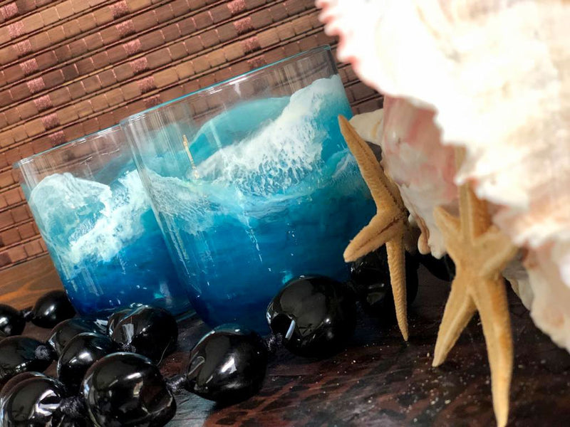 Pair of hand-painted short glass tumblers with blue, white, and aqua surfing waves, sitting among beach house decor.