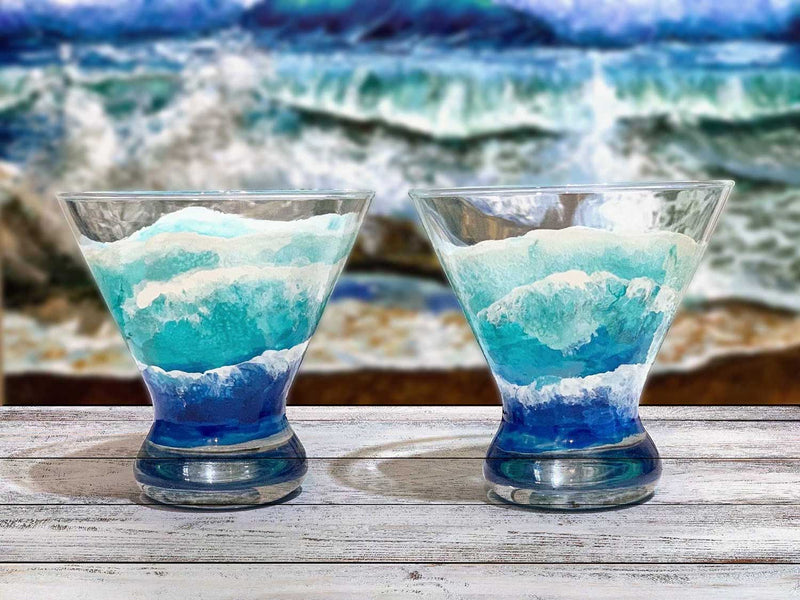 Pair of custom glasses for wedding, with blue ocean waves seen from birds-eye-view, sitting on a wood table in front of a beach oil painting of crashing waves.
