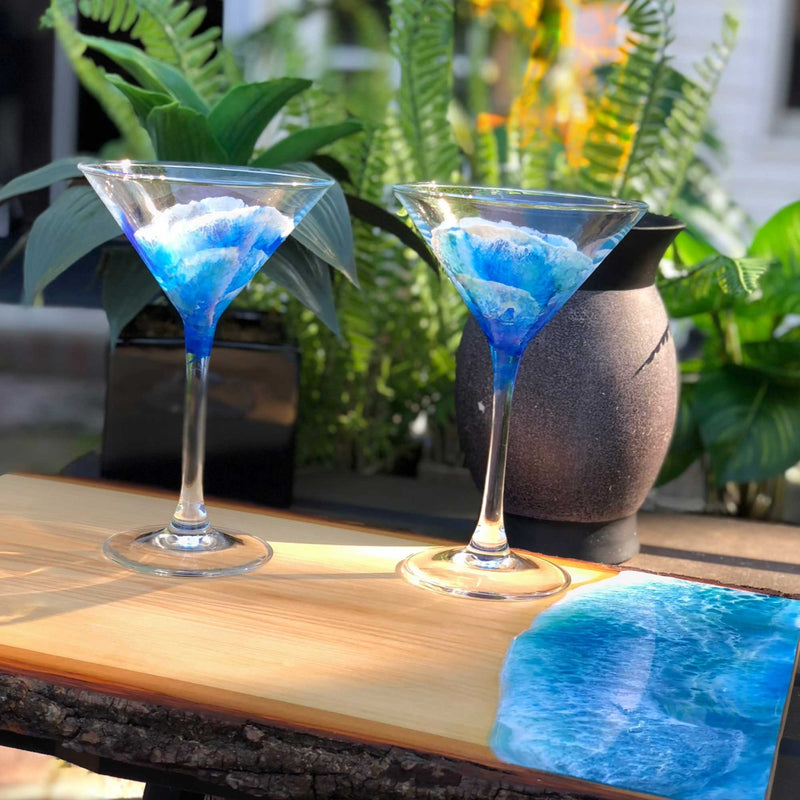 A pair of martini glasses hand painted with blues and whites to look like the ocean stand on a cutting board painted with art resin against a backdrop of trees and tiki torches