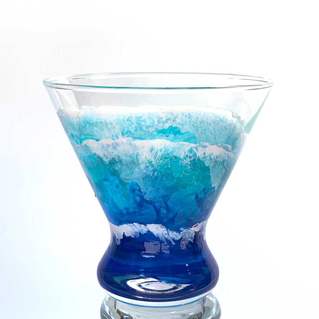 Custom cocktail glassware of hand-painted ocean waves washing up a stemless martini glass in blue, white, and turquoise.
