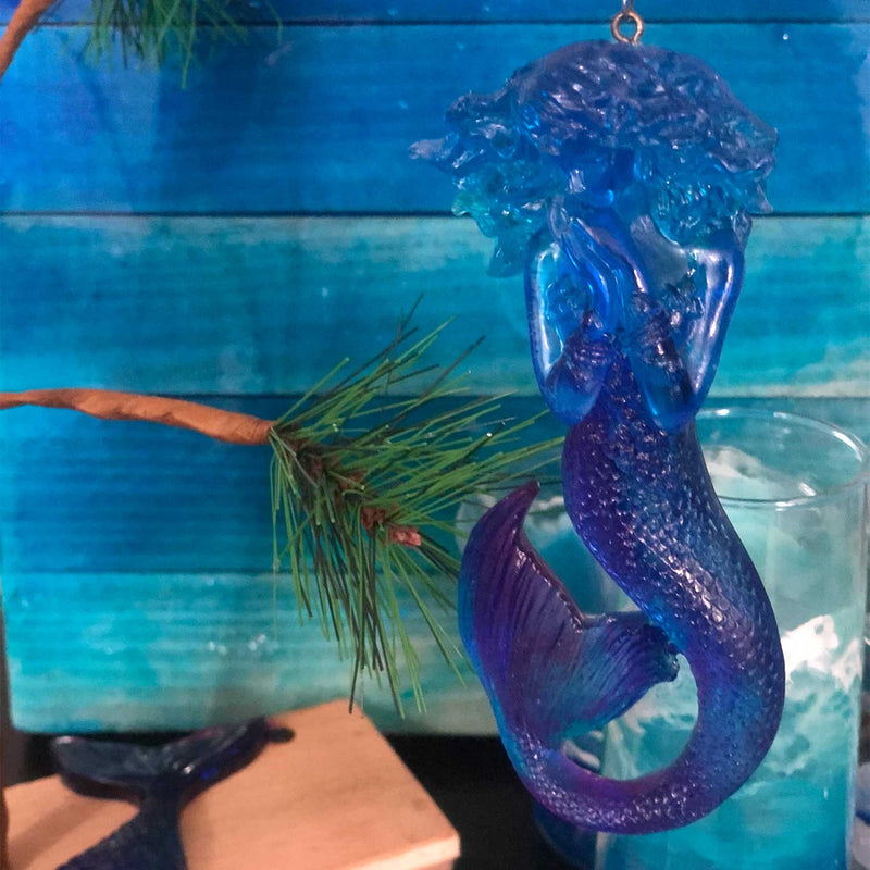 Blue mermaid Christmas tree ornament with purple accents, hanging from a small mermaid Christmas tree in front of a turquoise background