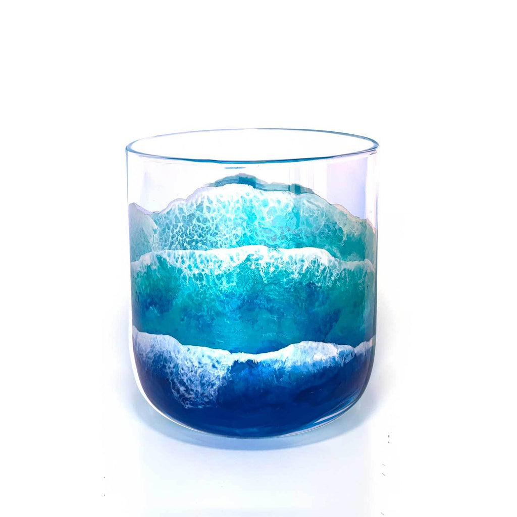 Short glass tumbler with blue and white ocean waves painted around the whole glass, as though washing up the beach.