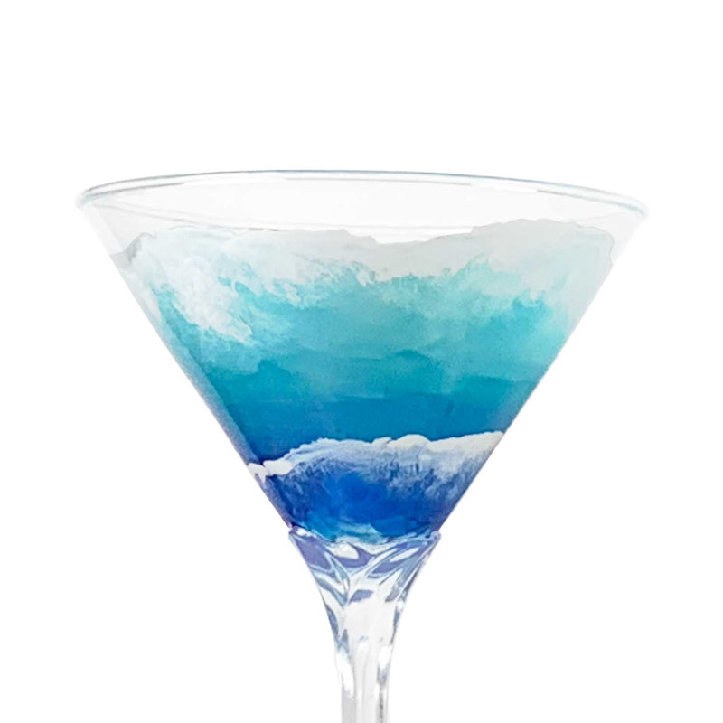 Close view of hand-painted martini glass with blue and white waves rolling up the beach. Luxury nautical barware for any occasion.