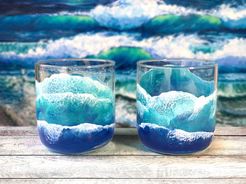 Pair of beach lover gifts, old fashioned glasses with hand-painted surfing waves washing up the sides. Sitting in front of an east coast beach painting. 