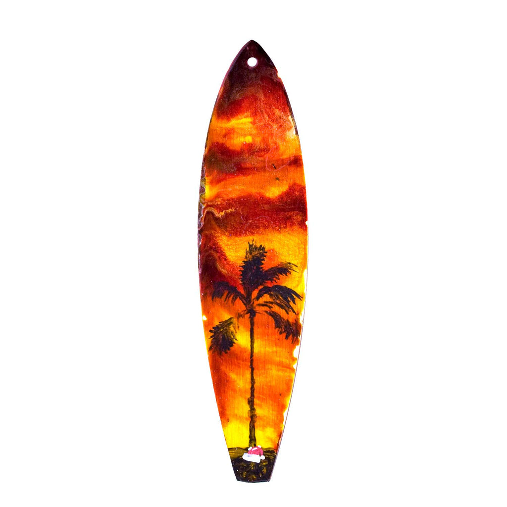 Wood surfboard christmas ornament with Santa hat on the beach beneath a palm tree silhouetted against an island sunset.
