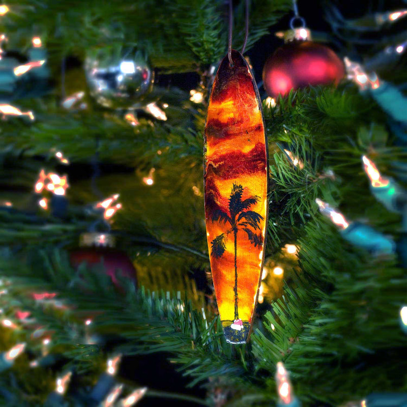 Coastal Christmas tree ornaments with surfing Santa hat sitting on the beach beneath a palm tree, with a tropical sunset.